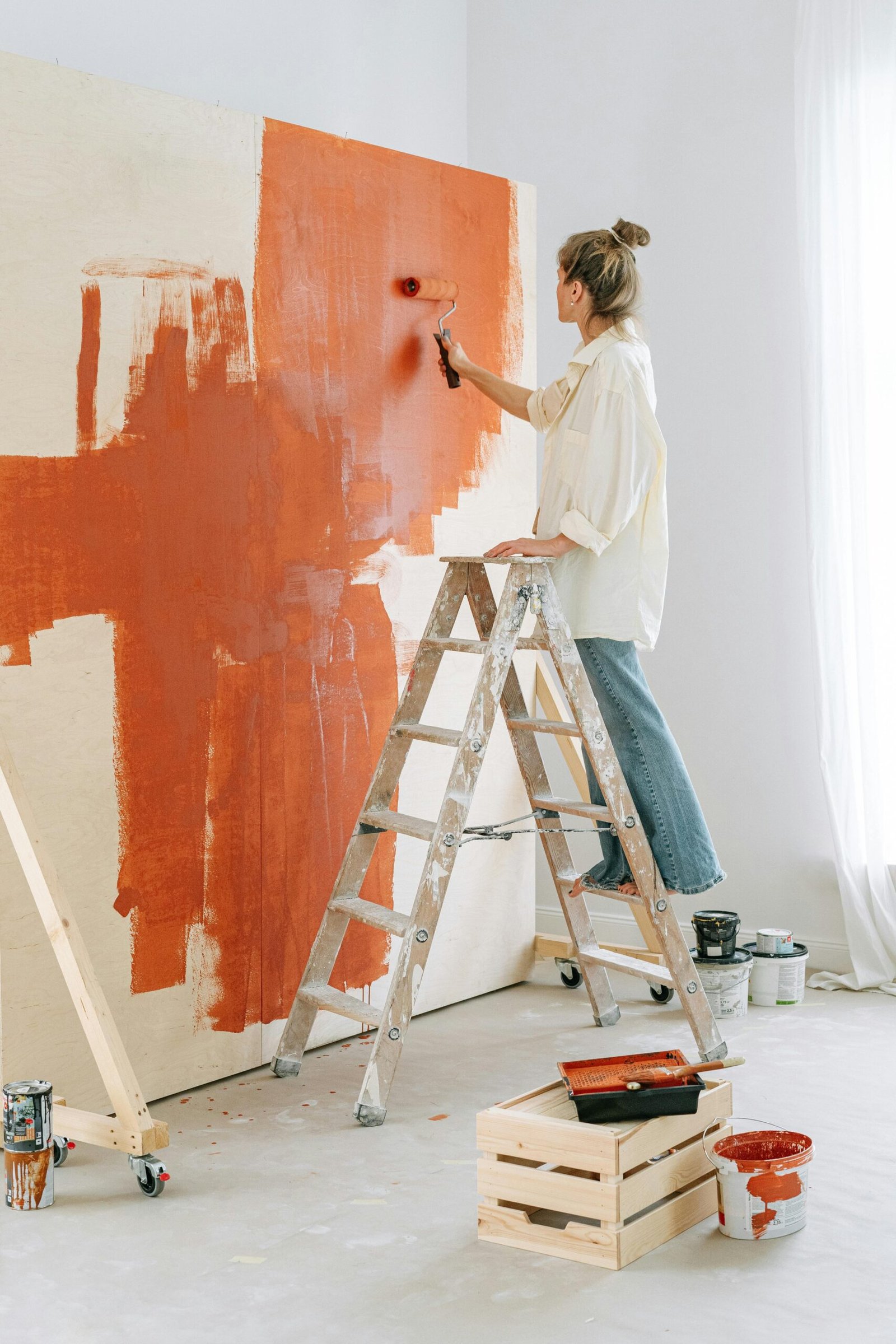 A woman stands on a stepladder, painting a room's wall during home renovation.